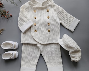 Puede incluir: Un conjunto de ropa de bebé de color crema, que incluye una chaqueta con botones de madera, pantalones a juego, una gorra y zapatos de bebé. El conjunto presenta un patrón texturizado y tejido. La chaqueta tiene un diseño cruzado. Los zapatos tienen una correa decorativa.