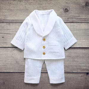May include: A white long-sleeved jacket and pants set featuring a textured chevron pattern. The jacket has a smooth shawl collar, three light brown buttons down the front, and a single chest pocket. The pants have plain white cuffs at the hem.