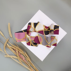 Puede incluir: Dos pañuelos de seda para el cabello con un estampado de eslabones de cadena en tonos rosa, negro, gris y dorado. Los pañuelos están colocados sobre un cuadrado blanco sobre un fondo gris, con espigas de trigo en la esquina inferior izquierda.