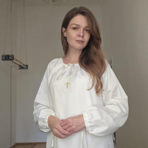 May include: A white tunic with long sleeves, a round neckline, and a gold cross embroidered on the chest. The tunic has a loose fit and gathered cuffs. The fabric looks like lightweight cotton or linen.