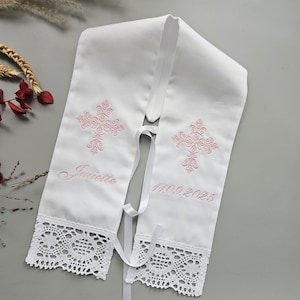 May include: White baptismal stole with pink embroidered crosses, the name "Juliette", and the date "11.09.2025". The stole has a white lace trim at the bottom and a ribbon tie.