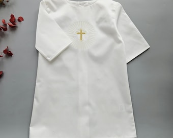 May include: A white, short-sleeved garment with a round neckline. The front features a gold cross embroidered within a sunburst design. The garment is likely for a religious ceremony, such as a baptism or first communion.