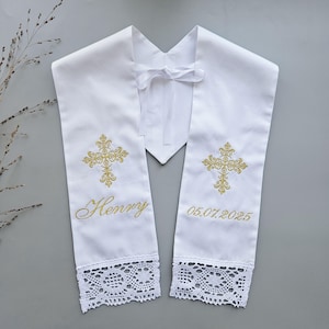 May include: White baptismal stole with gold embroidered cross design, the name "Henry" and the date "05.07.2025". The stole has a lace trim at the bottom and a white ribbon tie.