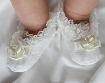 Könnte beinhalten: Ein Paar weiße Baby-Schühchen mit zarter Spitzenborte und einem Satinrosen-Detail. Die Schühchen sind aus einem weichen, strukturierten Stoff gefertigt und für Säuglinge konzipiert.