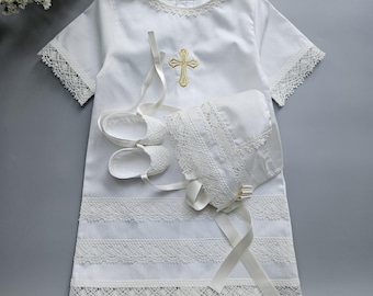 May include: A white short-sleeved dress featuring a gold embroidered cross on the chest, intricate lace trim on the sleeves and hem, and two horizontal lace bands. Included are matching white fabric booties and a bonnet with lace detailing.
