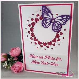 May include: A white card with a pink border and a purple butterfly cutout. The butterfly is centered on a circle of pink glitter hearts. The text "Hier ist Platz für Ihre Text-Idee" is printed on the card.