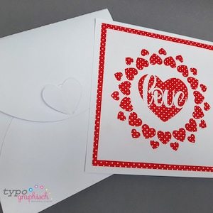 May include: A white card with a red heart and the word "love" inside a circle of red hearts. The card has a red and white polka dot border. The card is sitting on a white envelope with a heart cut out.