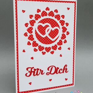 May include: A white card with a red border and red polka dots. The card has a red heart design with two hearts intertwined in the center. The text "Für Dich" is written in red on the card.