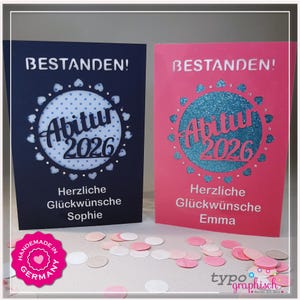 Puede incluir: Dos tarjetas de graduación, una azul marino y otra rosa, con el texto alemán "BESTANDEN!". Cada tarjeta presenta un diseño circular con "Abitur 2026" y un mensaje de felicitación. Las tarjetas están decoradas con detalles en forma de corazón.