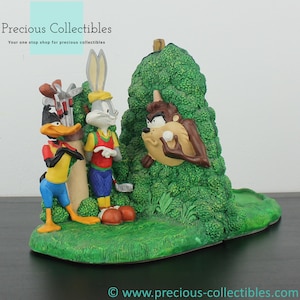 Puede incluir: Un sujetalibros con Bugs Bunny, Daffy Duck y Taz de Looney Tunes. Los personajes están representados jugando al golf sobre una base verde. Bugs Bunny lleva una camisa de golf roja y blanca, Daffy Duck lleva una camisa azul y pantalones amarillos, y Taz asoma desde detrás de un arbusto verde.