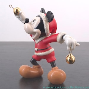 May include: A Mickey Mouse figurine dressed in a Santa Claus outfit, holding two gold Christmas ornaments. The figurine is wearing a red suit with white trim, a Santa hat, and brown shoes. The figure is a collectible item.