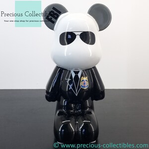 Peut inclure: Une figurine en céramique noire et blanche d'un ours portant un costume noir, des lunettes de soleil et un badge qui dit "FBI".