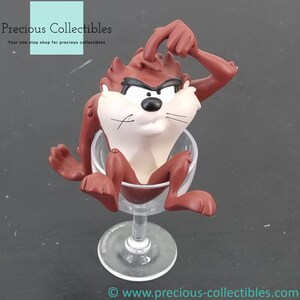Könnte beinhalten: Eine braune und weiße Cartoon-Figur des Tasmanian Devils, die in einem klaren Weinglas aus Glas sitzt. Der Teufel hat einen schelmischen Ausdruck und einen Finger auf dem Kopf, als würde er nachdenken.