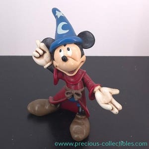 Puede incluir: Figura de Mickey Mouse vestido de hechicero, con un sombrero azul con una estrella y una luna creciente blancas, una túnica roja y zapatos marrones. Una mano levantada y la otra extendida, en pose mágica.