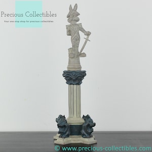 Puede incluir: Una figurita de cerámica blanca de un conejo vestido de soldado de pie sobre una columna con una base azul y blanca. El conejo sostiene una espada y lleva un sombrero azul con una pluma. La base de la columna tiene cuatro cabezas de león.