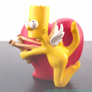 May include: A yellow Bart Simpson figurine in a Cupid pose, with white wings and a bow and arrow, positioned in front of a red heart. This collectible figurine is likely made of resin or a similar material.
