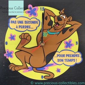 Puede incluir: Un reloj de pared amarillo y marrón con Scooby Doo de la caricatura Scooby Doo. El reloj tiene un fondo negro y las palabras "PAS UNE SECONDE À PERDRE... ...POUR PRENDRE SON TEMPS!" en un globo de diálogo.