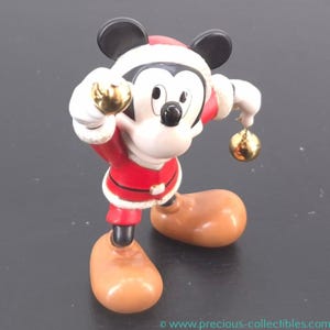 May include: A Mickey Mouse figurine dressed in a Santa Claus outfit, holding two golden ornaments. The figurine has a red and white Santa suit, brown shoes, and black ears. The figurine is a collectible item.