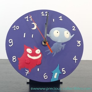 May include: A purple clock with a red and white cat, a blue ghost, and a white house on the face. The clock hands are black and red.