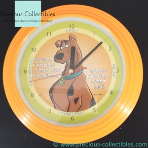 Puede incluir: Un reloj de pared amarillo y verde con un personaje de dibujos animados de Scooby Doo. La esfera del reloj tiene el texto "COOL!" en la parte superior, "DEUX MINUTES!" a la izquierda, "Y'A PAS L'FEU!" en la parte inferior y "RELAX!" a la derecha. Las manecillas del reloj apuntan a las 12 y las 3.