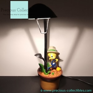 Könnte beinhalten: Eine gelbe Vogelfigur, die einen Hut trägt und eine Gießkanne hält, sitzt in einem braunen Topf mit grünen Pflanzen. Die Figur ist Teil einer Schreibtischlampe mit einem schwarzen Metallfuß und einem schwarzen Lampenschirm.