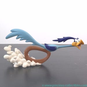 May include: A figurine of the Road Runner cartoon character in mid-run. The bird is blue, with a yellow beak and open mouth. A brown loop and white cloud-like shapes are behind the bird, suggesting movement. The figurine is on a dark surface.