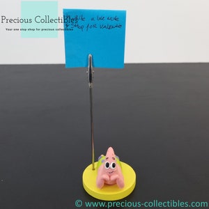 Puede incluir: Una figurita de personaje de dibujos animados rosa con una base amarilla y una tarjeta de nota azul con el texto "Website a late male Shop for Valentine" adjunta a un soporte plateado.