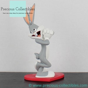 Puede incluir: Una figurita de Bugs Bunny gris con una expresión de sorpresa, de pie sobre una base roja en forma de corazón. La figurita forma parte de una colección de personajes de dibujos animados.