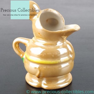 Peut inclure: Un petit pichet en c&eacute;ramique brun avec une bande verte et jaune autour du milieu. Le pichet a une base large et ronde et un bec qui s'incurve vers le haut et vers l'ext&eacute;rieur.