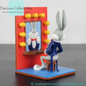 Puede incluir: Una figura de Bugs Bunny sentado en un taburete, frente a un espejo con su reflejo. La escena está ambientada sobre un fondo rojo con detalles amarillos. La figura lleva un traje azul y una pajarita roja. La base es azul.