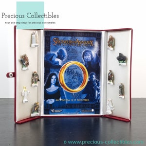 Op de afbeelding: Een rode displaykast met een wit interieur, waarin een verzameling Lord of the Rings-figuurtjes wordt getoond. De kast is open en laat een poster zien voor de film "The Lord of the Rings: The Return of the King" met de tekst "Au cinéma le 17 décembre".