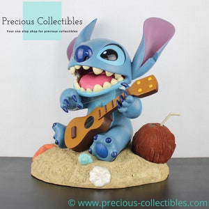 Puede incluir: Una figura azul de Stitch tocando un ukelele, sentada en una playa de arena con un coco y conchas. La figura forma parte de una colecci&oacute;n de Disney.