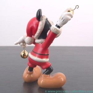 May include: A ceramic Mickey Mouse figurine dressed in a Santa Claus outfit, holding a gold ornament. The figurine is red, black, and white with gold accents. The figure is posed with one arm raised, holding a gold ornament.