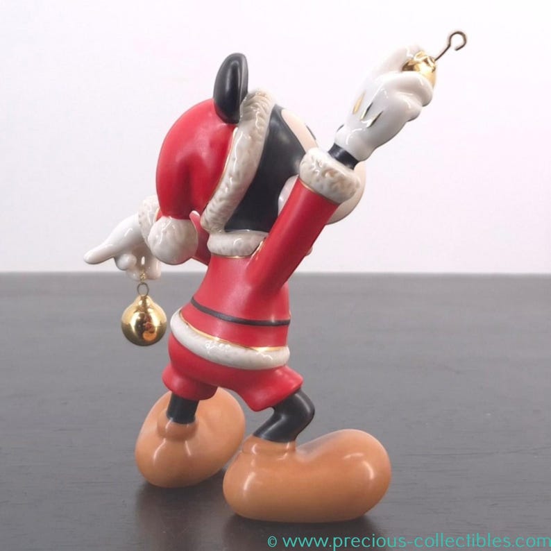 May include: A ceramic Mickey Mouse figurine dressed in a Santa Claus outfit, holding a gold ornament. The figurine is red, black, and white with gold accents. The figure is posed with one arm raised, holding a gold ornament.