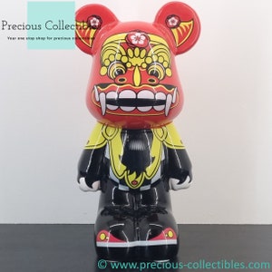May include: Red, yellow, and black bear figurine with a lion's face. The bear is wearing a yellow and black patterned vest. The bear's face is red with a white mouth and teeth. The bear's eyes are black and the bear's nose is white.