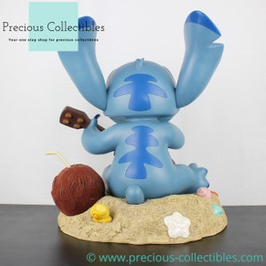 Puede incluir: Figurilla azul de Stitch tocando un ukelele, sentada en una escena de playa con un coco, conchas y una estrella de mar. La figurilla es parte de una colecci&oacute;n de Disney.