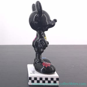 May include: A glossy black figurine of a cartoon character with large ears, standing on a white base with a black and white checkered pattern. The figurine has a red floral design on its foot and a pink heart on its chest.