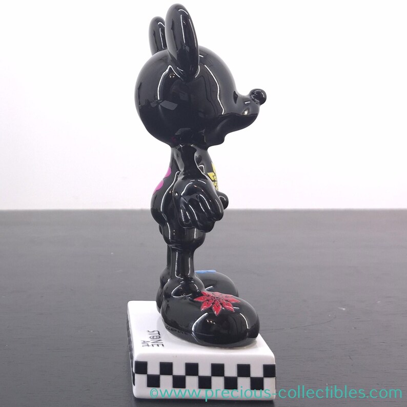 May include: A glossy black figurine of a cartoon character with large ears, standing on a white base with a black and white checkered pattern. The figurine has a red floral design on its foot and a pink heart on its chest.