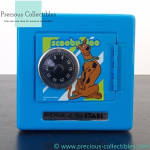 Puede incluir: Hucha de metal azul con forma de caja fuerte con un diseño de Scooby Doo. La caja fuerte tiene una cerradura de combinación y una puerta que se abre. La parte delantera de la caja fuerte presenta una imagen de dibujos animados de Scooby Doo con el texto "Scooby Doo" y "Avenue of the Stars".
