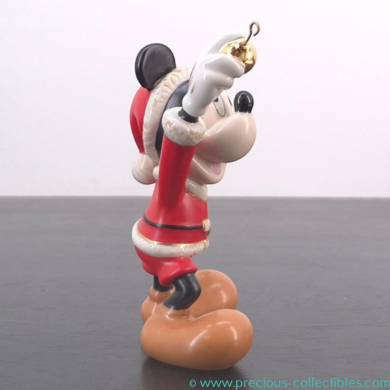 May include: A ceramic Mickey Mouse Christmas ornament. Mickey is dressed in a red Santa suit with white trim, holding a gold ornament. The ornament has black ears, a white face, and large tan feet.