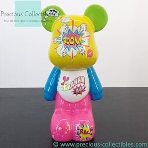 Puede incluir: Una figurita de oso de cerámica amarilla, rosa, azul y verde con un diseño de cómic. El oso tiene una cara blanca con una marca de beso rosa y las palabras "BOOM" y "MIAOW" en una fuente de estilo cómic. El oso lleva una camisa azul con corazones rosas y una falda rosa con estrellas y la palabra "CRACK" en una fuente de estilo cómic.