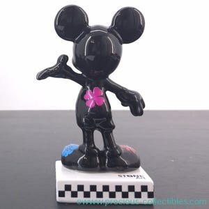 May include: A black Mickey Mouse figurine with a glossy finish. The figurine has a pink flower on its chest and colorful accents on its feet. It stands on a white base with a black and white checkered pattern. The base reads "STONE ART".