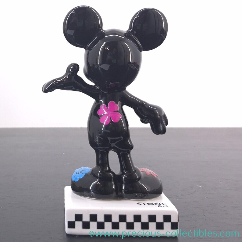 May include: A black Mickey Mouse figurine with a glossy finish. The figurine has a pink flower on its chest and colorful accents on its feet. It stands on a white base with a black and white checkered pattern. The base reads "STONE ART".