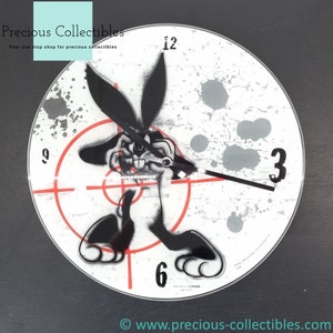 Puede incluir: Un reloj blanco y negro con un diseño de diana y un conejo de dibujos animados en el centro. El conejo lleva un collar de perlas y tiene una expresión traviesa. La esfera del reloj tiene números y manecillas negros.
