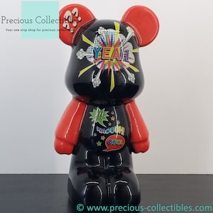 Peut inclure: Figurines d'ours de dessin animé noir et rouge avec une finition brillante. L'ours a une bulle de dialogue sur la tête qui dit "YEAH!" et une autre bulle de dialogue sur la poitrine qui dit "COOL!" L'ours a également une bulle de dialogue sur le ventre qui dit "WACOUHI!"