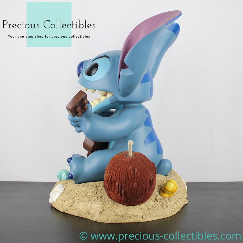 Puede incluir: Una figura azul de Stitch tocando un ukelele, sentada en una playa de arena con un coco marr&oacute;n y una concha amarilla. La figura forma parte de una colecci&oacute;n de Disney.