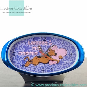 Puede incluir: Un despertador ovalado azul con un diseño floral morado y blanco. El reloj presenta una imagen de dibujos animados de Scooby Doo sosteniendo una almohada rosa en forma de corazón. La esfera del reloj dice "Laissez mo dormir" y tiene una manecilla de hora roja y una manecilla de minutos negra.