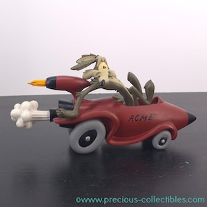 May include: A figurine of Wile E. Coyote in a red ACME car with a rocket engine. The coyote is olive green and the car has grey wheels. The rocket engine emits a cloud of white smoke and a yellow flame.