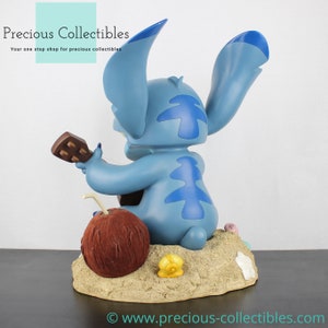 Puede incluir: Figurilla azul de Stitch tocando un ukelele, sentado en una playa de arena con un coco y conchas marinas. La figurilla es parte de una colecci&oacute;n de Disney.