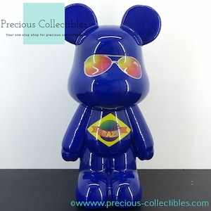 Peut inclure: Une figurine d'ours de dessin animé bleu brillant portant des lunettes de soleil arc-en-ciel et un t-shirt jaune avec le mot "BRAZIL" dessus.